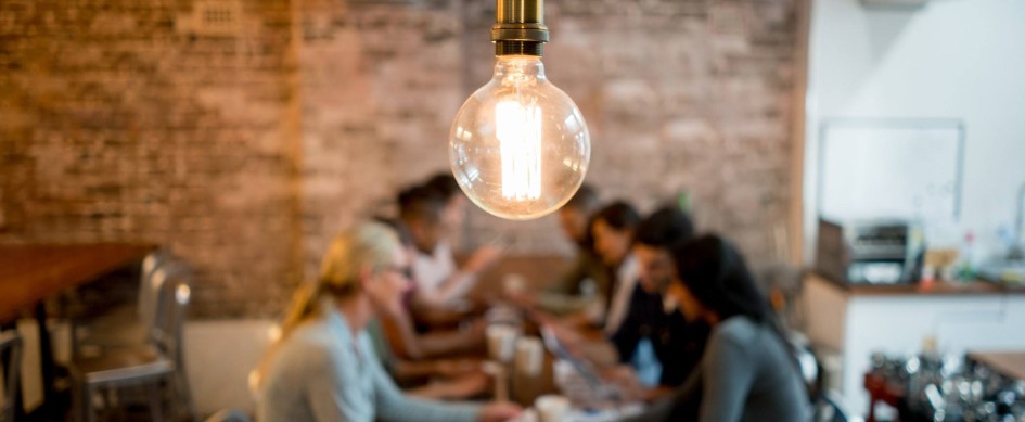 A Lightbulb, With A Team Around A Table In The Background